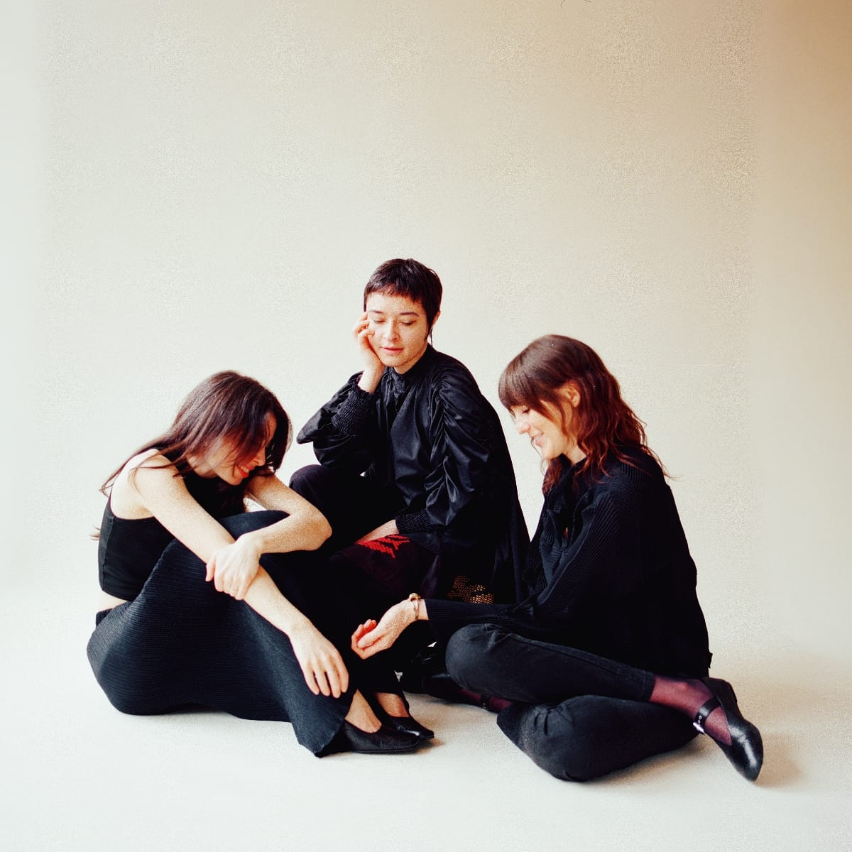 Whitney Johnson, Macie Stewart, and Lia Kohl sit clustered on a white floor in all-black clothing, heads tilted downward in a quiet, inward-facing pose. Photo by Leah Wendzinski.