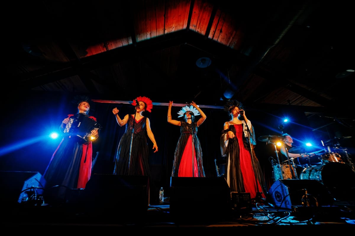 YAGÓDY perform at Big Ears 2026, four members in red and black gowns with floral headdresses singing beneath dramatic blue stage lights in a wood-beamed venue. Photo by Billie Wheeler.