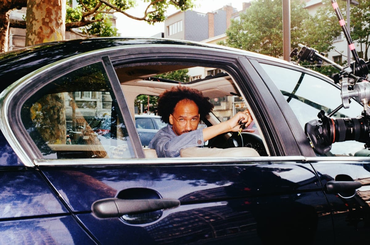 Youniss sits in the driver's seat of a dark blue car, looking back through the open window, with a film camera rig mounted to the exterior of the vehicle. Photo by Adel Setta.