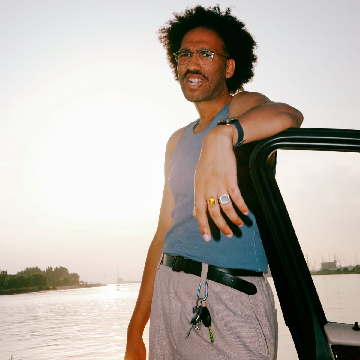Youniss leans against a car door in a blue tank top, rings on his fingers, with a sun-bleached river and industrial skyline behind him. Photo by Adel Setta.