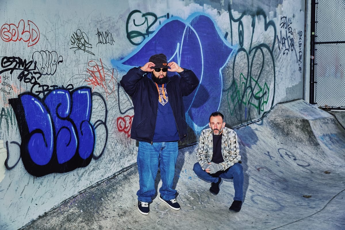 Dälek pose at a graffiti-covered skate park, one standing and adjusting his sunglasses, the other crouching on a concrete ramp. Photo by Jonny Scala.
