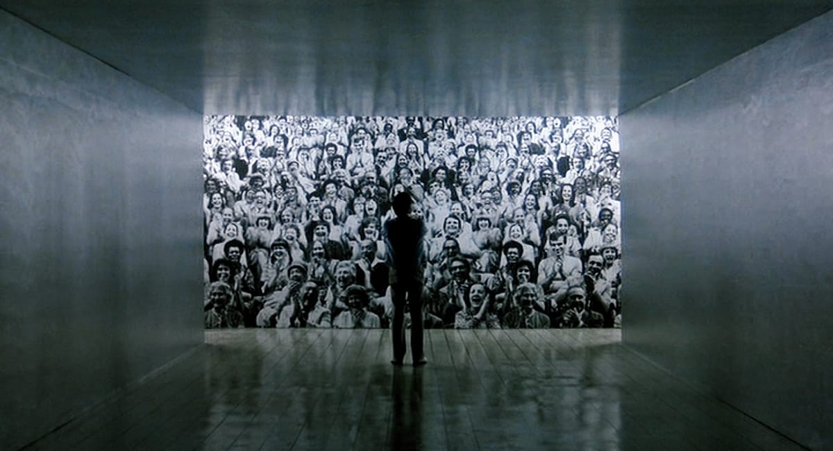 A silhouetted figure stands alone in a bare room before a giant projected image of a cheering crowd, in a still from The King of Comedy.