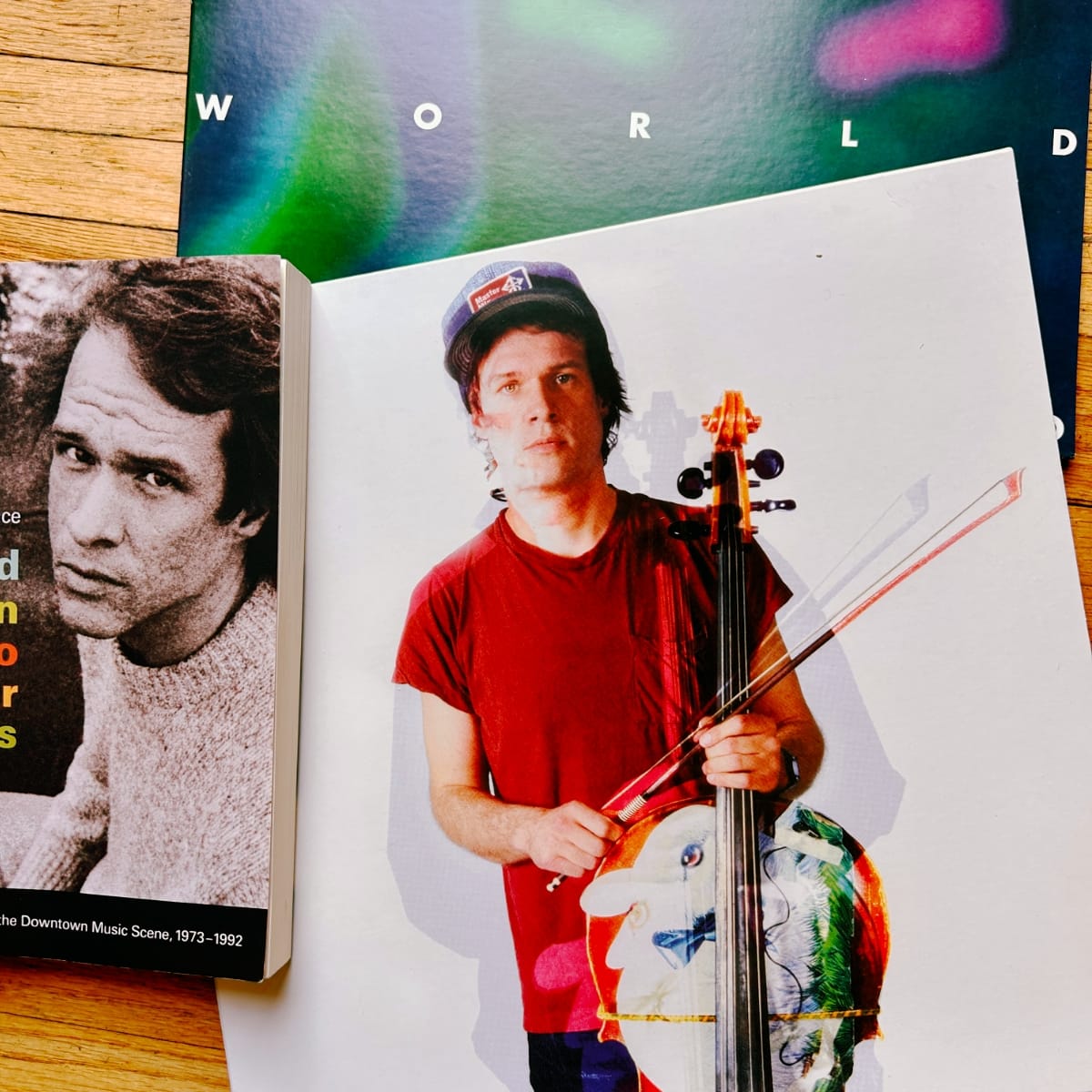 Open magazine spread showing a cellist holding instrument and bow, wearing a red t-shirt and cap. Beside it, a paperback book about the Downtown Music Scene, 1973–1992, with Arthur Russell on the cover.