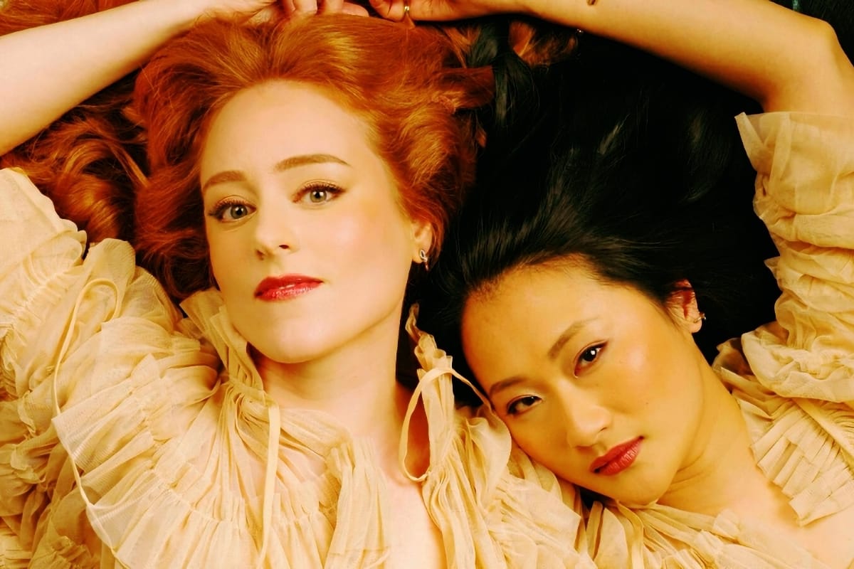Hannah Peel and Beibei Wang lie on their backs in matching cream ruffled gowns, hair fanned out around them, gazing upward at the camera against a pale blue-green background. Photo by Raphael Neal.