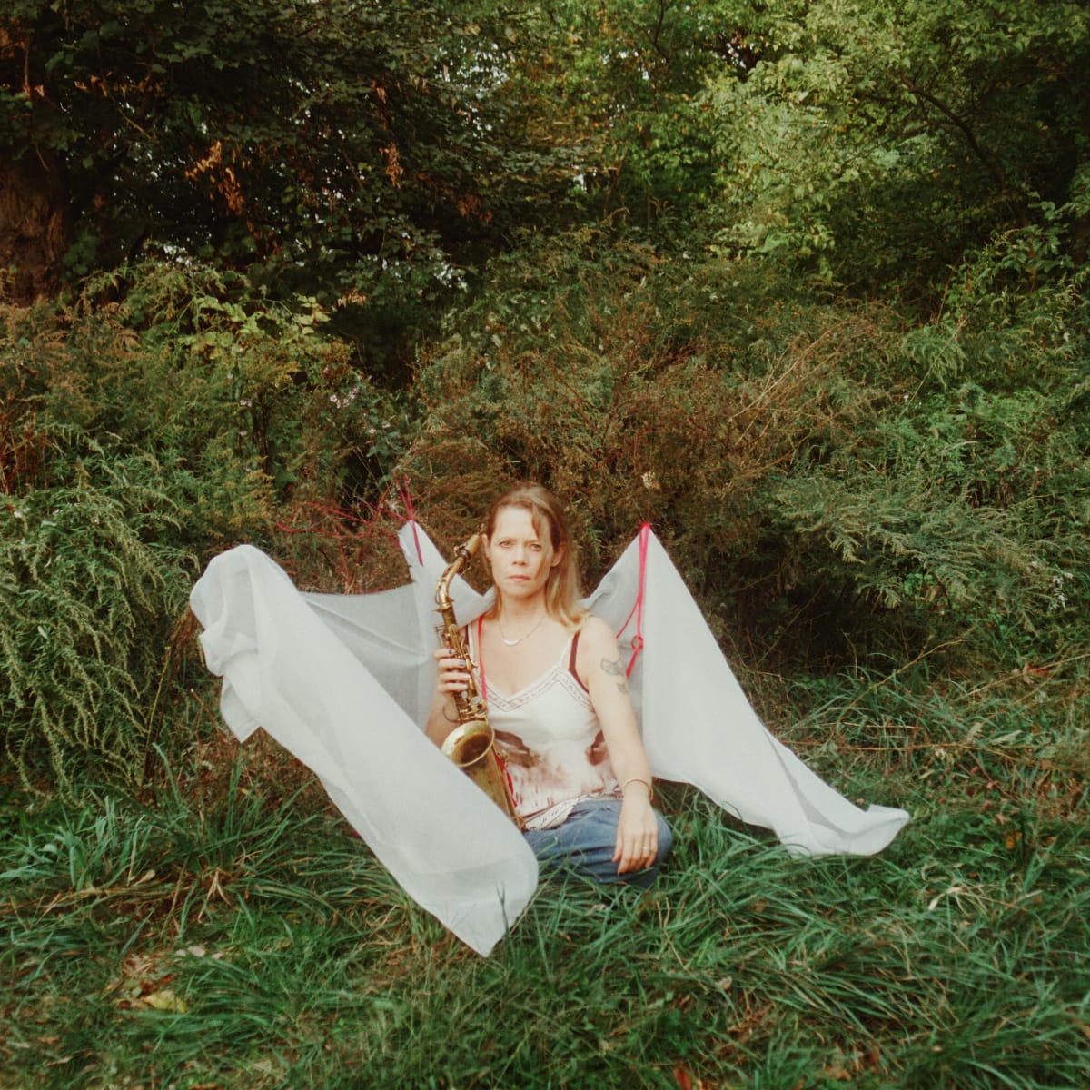 Caroline Davis sits in overgrown grass holding a saxophone, surrounded by billowing white fabric tied with red cord, dense green foliage behind her. Photo by Adi Meyerson.