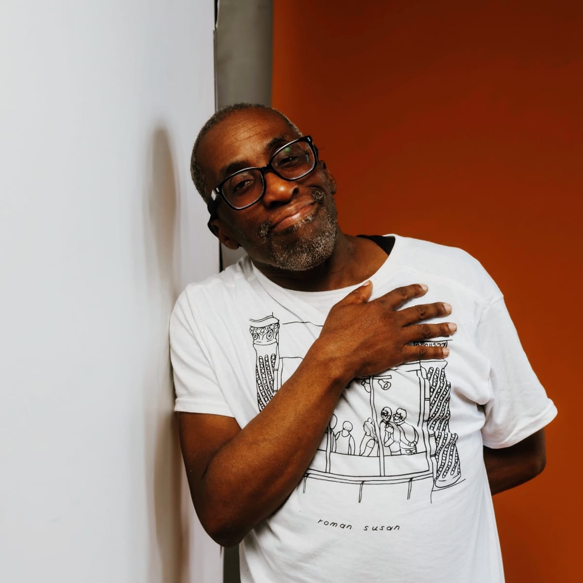 Charles Joseph Smith leans against a white wall, hand on chest, wearing a white graphic tee and dark-framed glasses, smiling slightly. Photo by Dennis Larance.