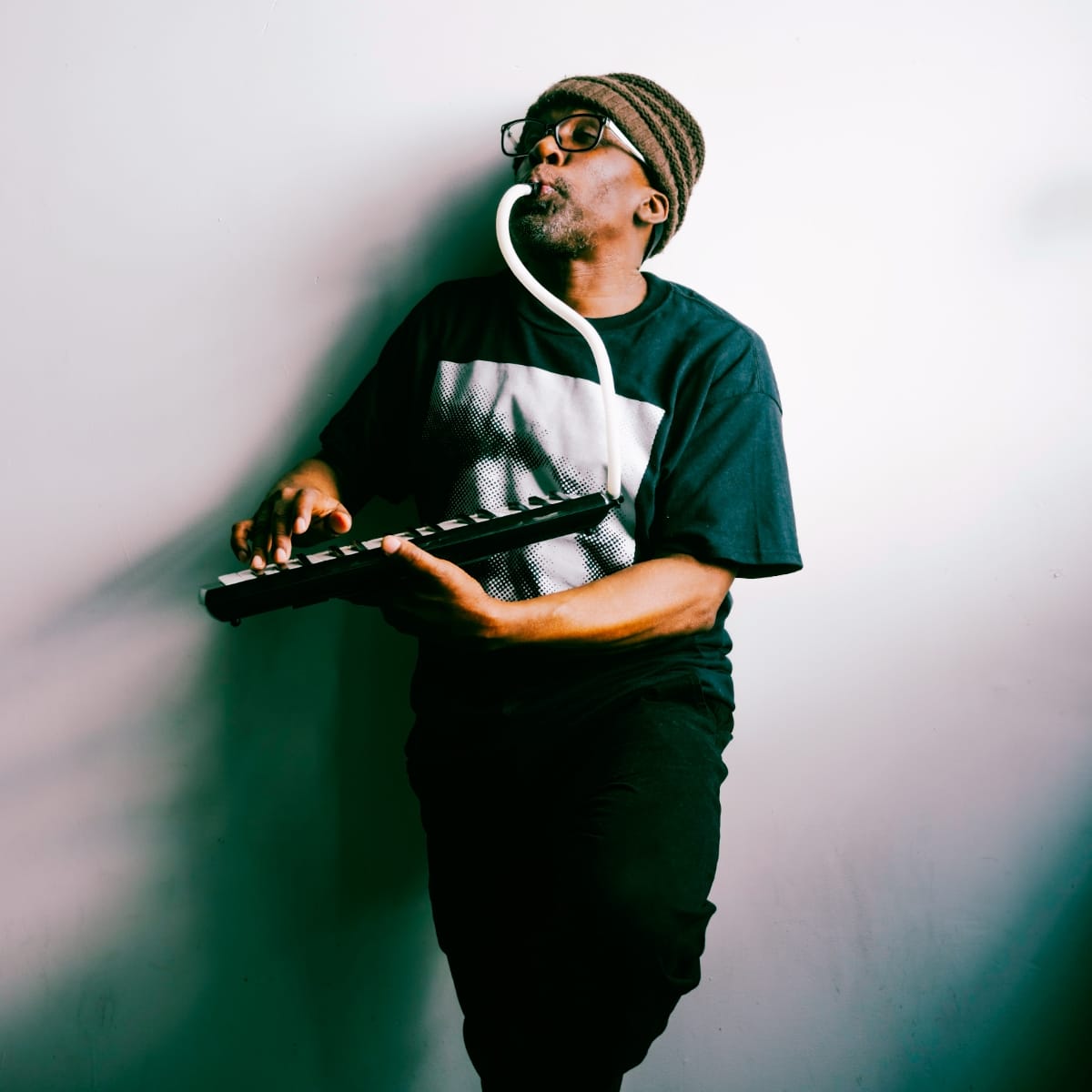 Charles Joseph Smith plays a keytar while blowing through a small wind instrument, wearing a knit beanie and glasses, against a softly lit pale wall. Photo by Dennis Larance.