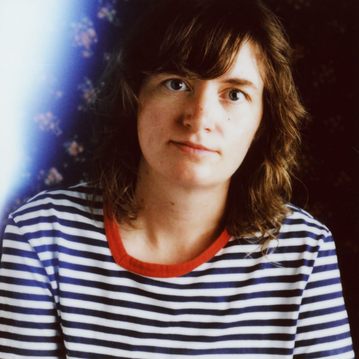Charlotte Cornfield in a blue-and-white striped shirt with a red collar, looking at the camera, shoulder-length wavy brown hair, against a dark background with a bright light bloom at left.
