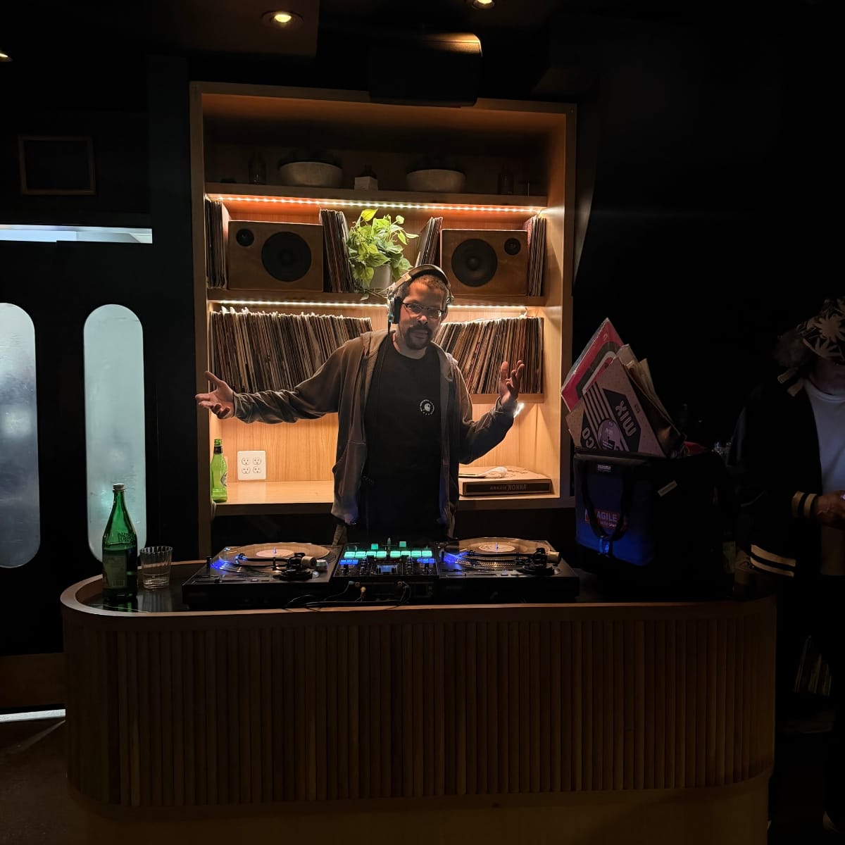 DJ Amir stands behind a vinyl DJ setup, arms open mid-gesture, headphones on, backlit by a shelving unit filled with records, speakers, and warm LED strips.
