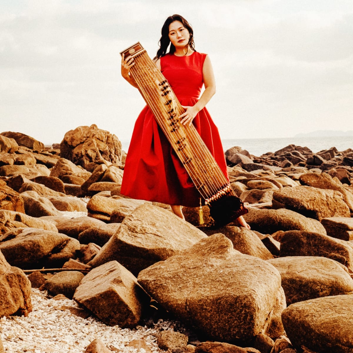DoYeon Kim stands barefoot on a rocky shoreline in a red dress, holding a gayageum upright, with the sea visible in the distance.