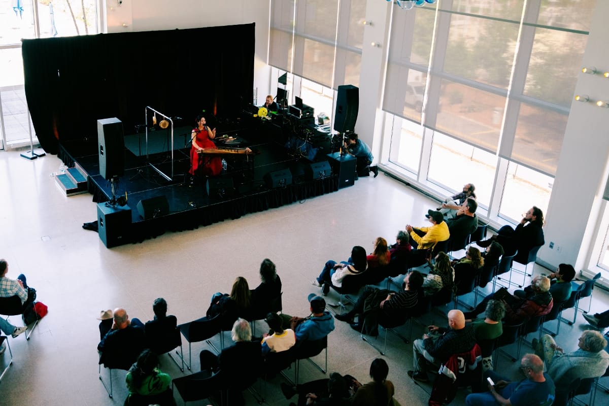 DoYeon Kim performs on gayageum in a red dress on a low stage in a sunlit gallery space, photographed from above with a seated audience looking on. Photo by Jess Maples.