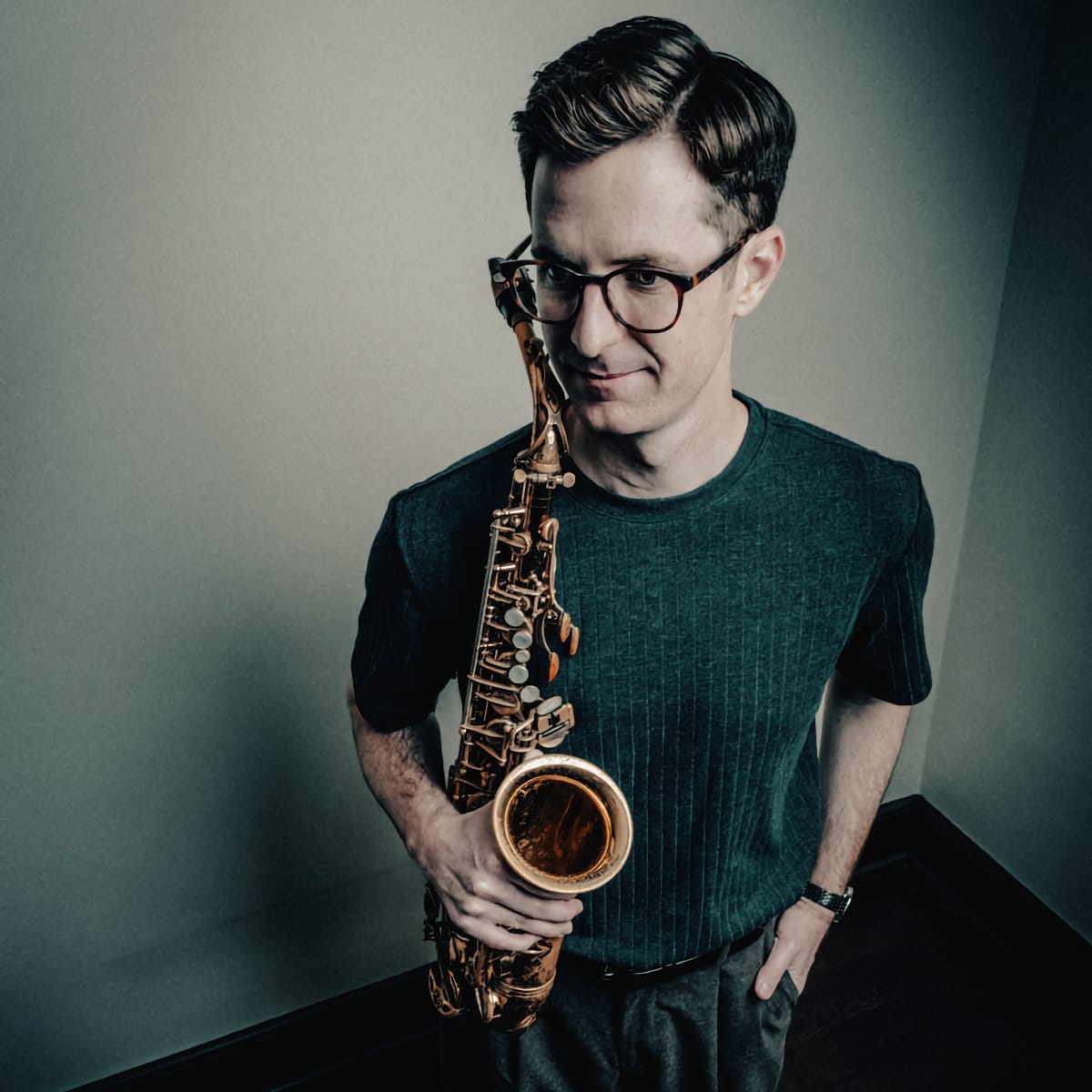 Ethan Helm stands holding a saxophone at his side, looking down toward the camera with a slight smile. Shot from above against a pale corner wall.