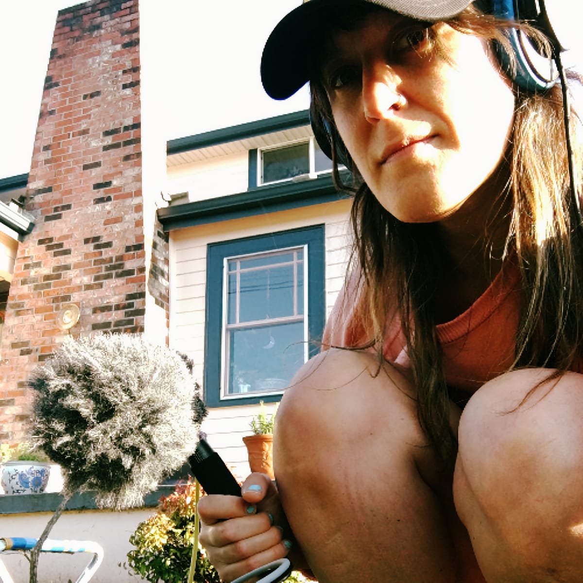Lisa Ann Schonberg holds a boom microphone with a furry windscreen outdoors, looking over her shoulder at camera. Baseball cap, headphones around neck. Brick chimney and house behind her.