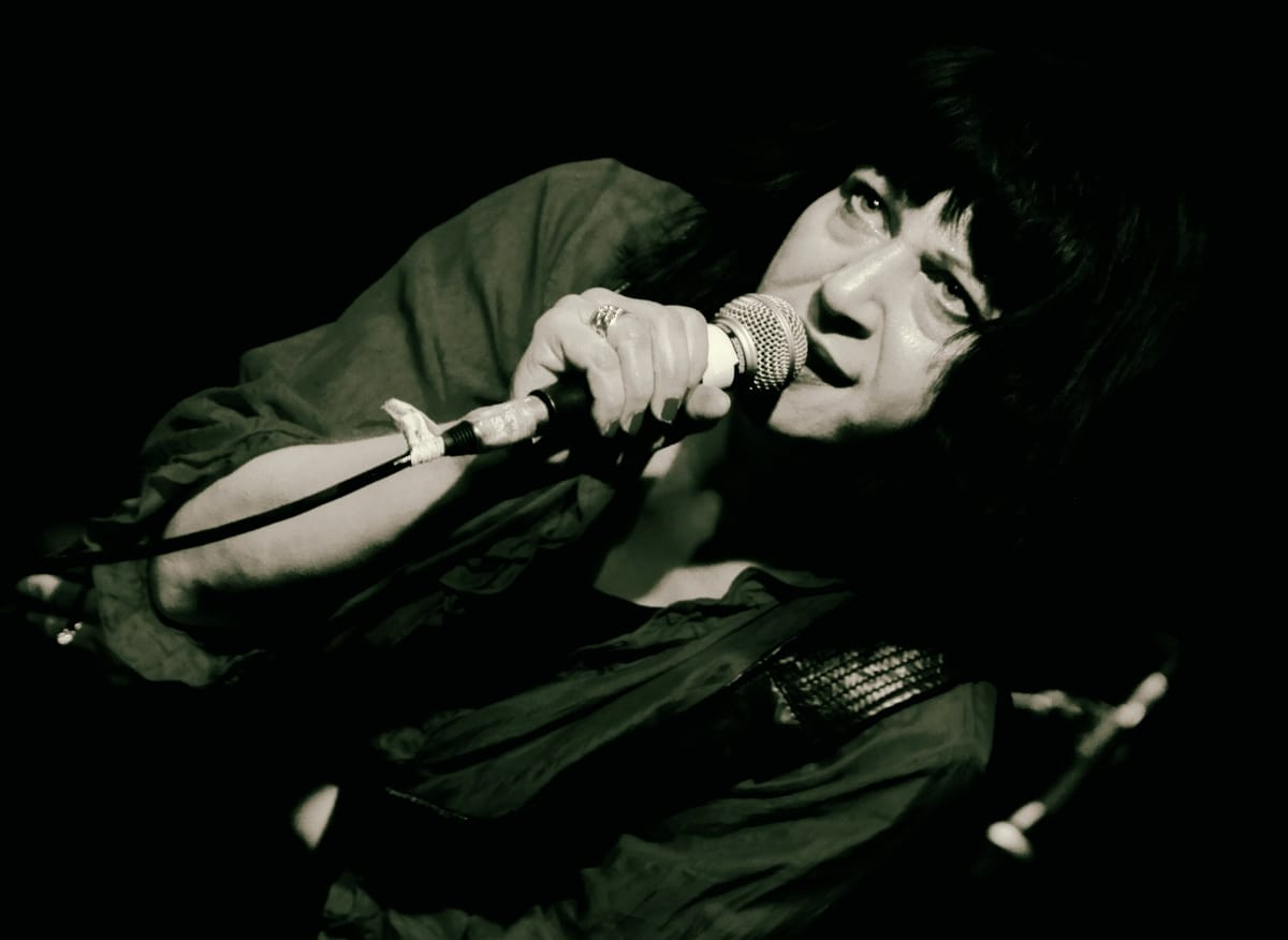 Lydia Lunch leans toward the mic, lips parted mid-performance, in a high-contrast black-and-white live photo shot from a low angle against a dark background.