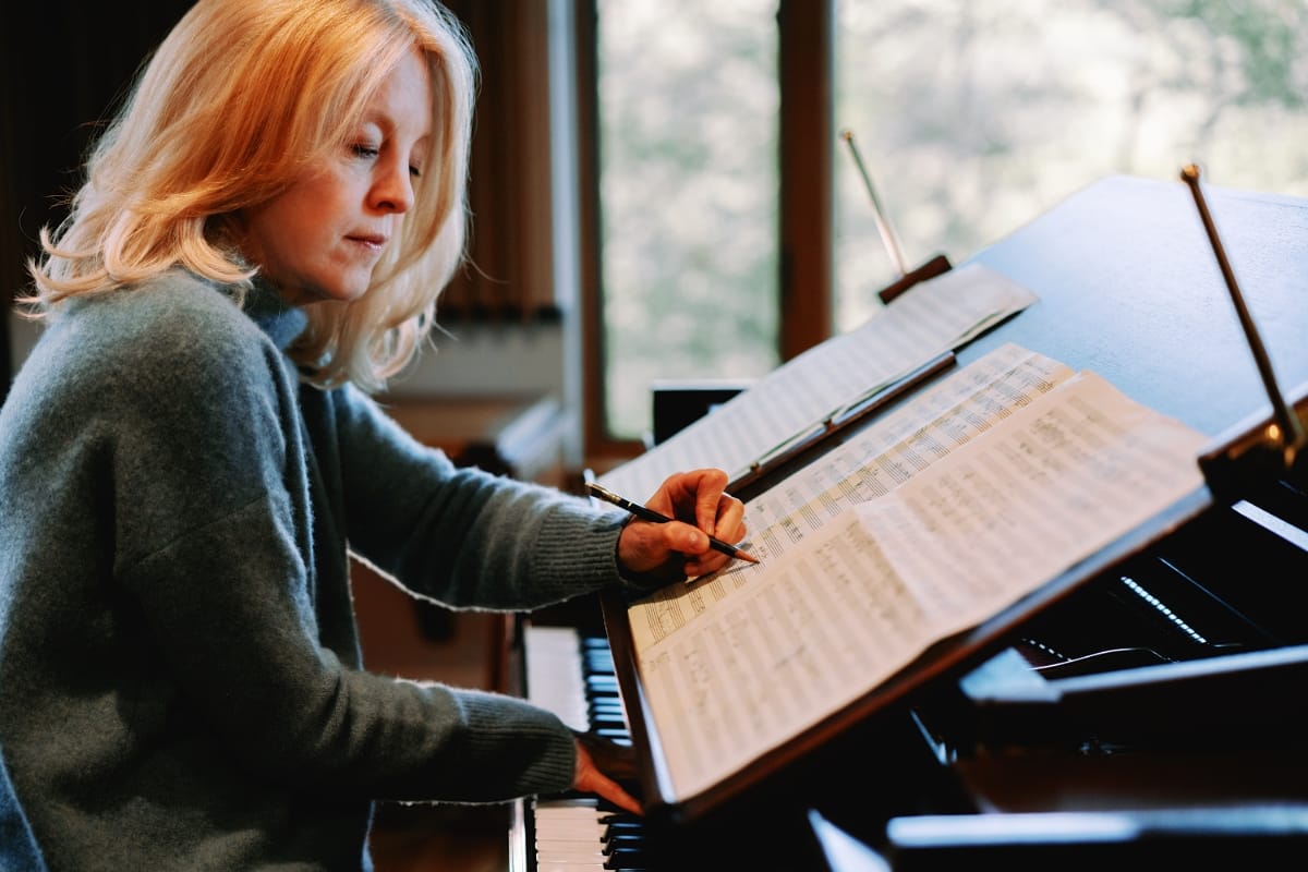 Maria Schneider leans over a grand piano, pencil in hand, marking up a full orchestral score. Photo by Briene Lermitte.