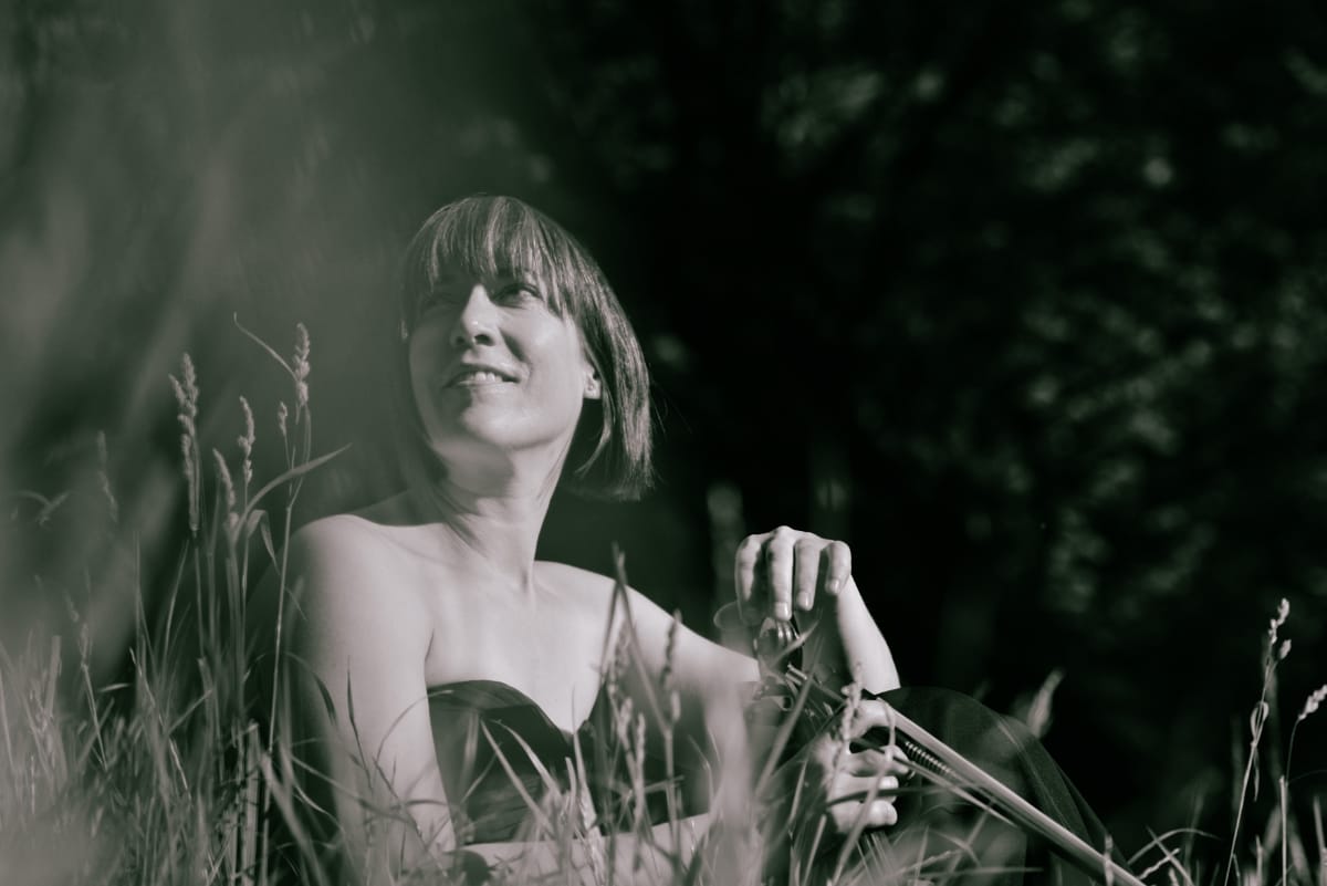 Meredith Bates sits among tall grasses outdoors, holding a violin and bow, glancing upward with a slight smile. Black-and-white portrait.