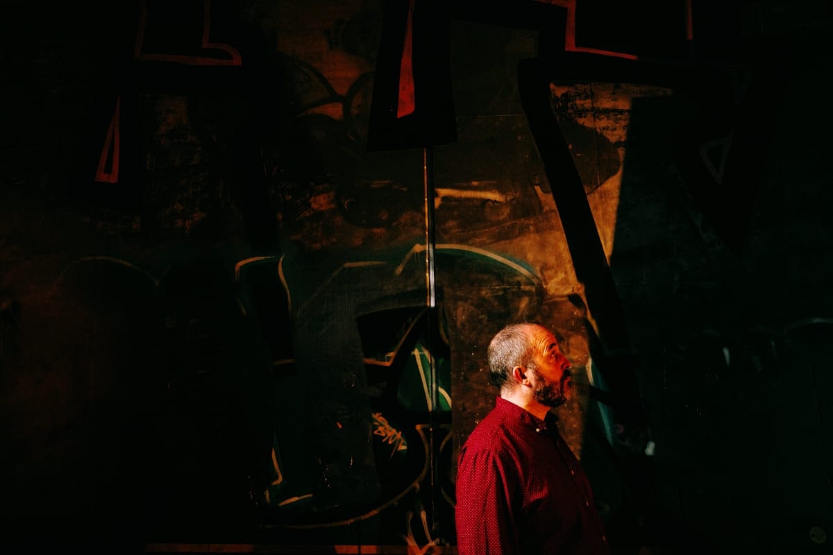 Nick Fraser, shown in profile from the shoulders up, gazes upward against a vast, darkened graffiti wall. A narrow shaft of light illuminates his face and red shirt against the surrounding darkness.