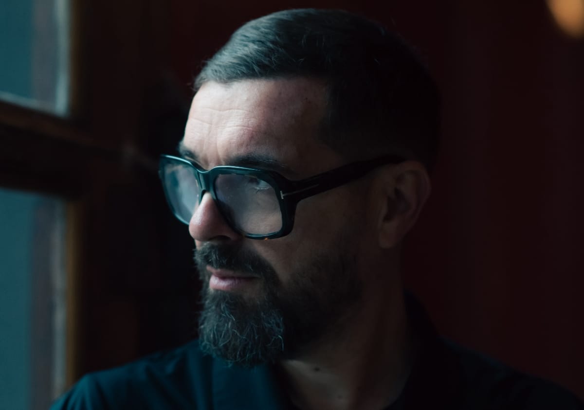 Close-up portrait of Speedy J in profile, wearing large square-framed tinted glasses, against a dark warm-toned background. Photo by Tommy Spring.