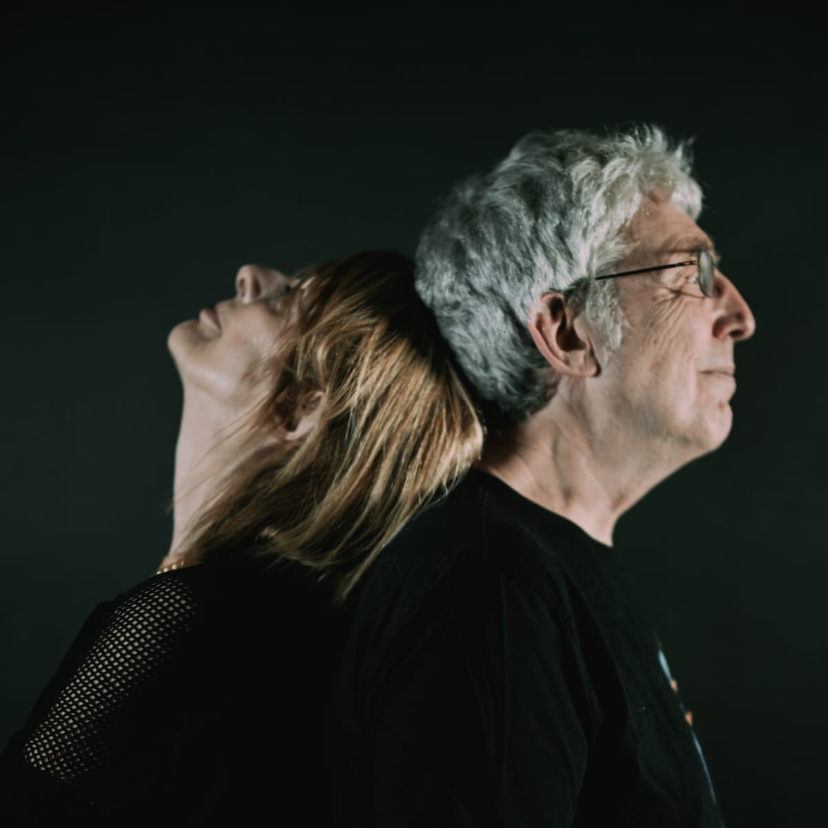 Miquette Giraudy and Steve Hillage of System 7 stand back-to-back against a dark background, both in profile, each facing away from the other.