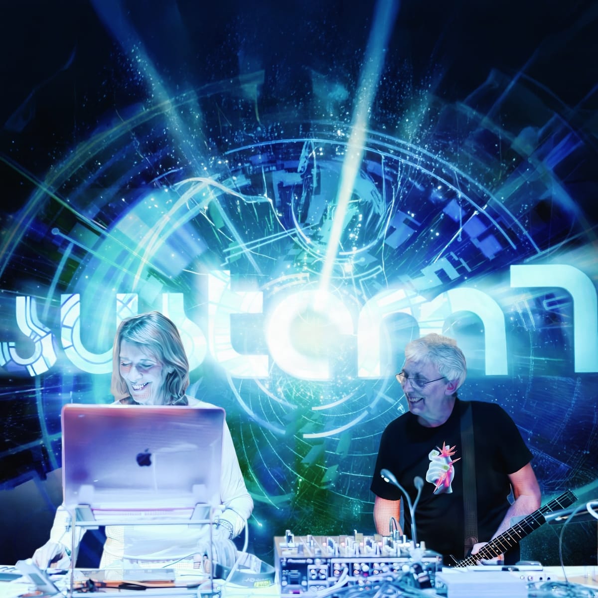 Miquette Giraudy works a laptop and mixer while Steve Hillage plays guitar beside her, both smiling mid-performance against a vivid blue-green digital projection backdrop.
