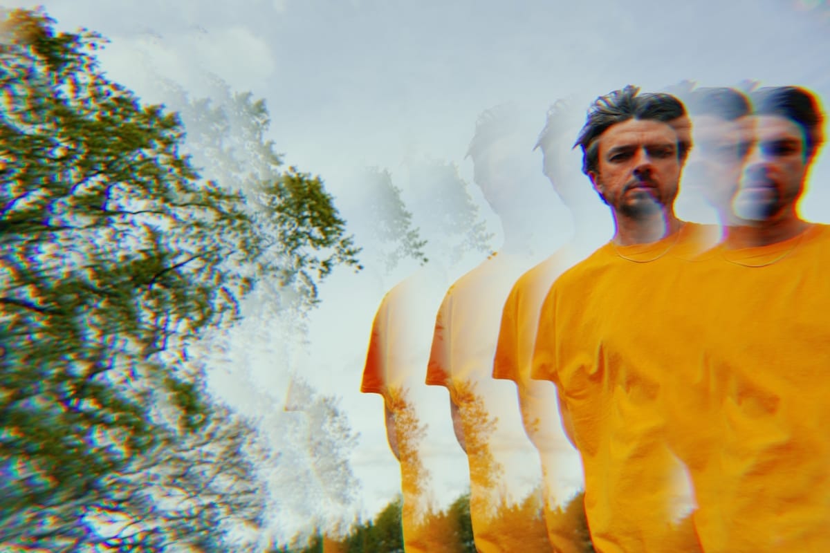 Tom Skinner in a yellow t-shirt and chain necklace, multiplied by a prism effect into a receding row of overlapping figures against an overcast sky and tree line. Photo by Jason Evans,