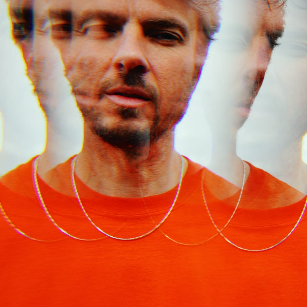 Tom Skinner in a red-orange t-shirt and chain necklace, shot in close-up with a prism effect that fractures his image into overlapping exposures against a bright, blown-out background. Photo by Jason Evans.
