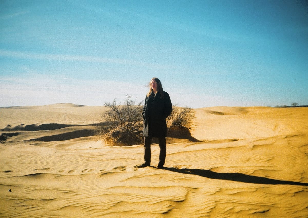 Johnathon Ford of Unwed Sailor stands on a sand dune in a dark coat, long hair wind-blown, a bare desert shrub behind him under a clear blue sky. Photo by Charles Elmore.