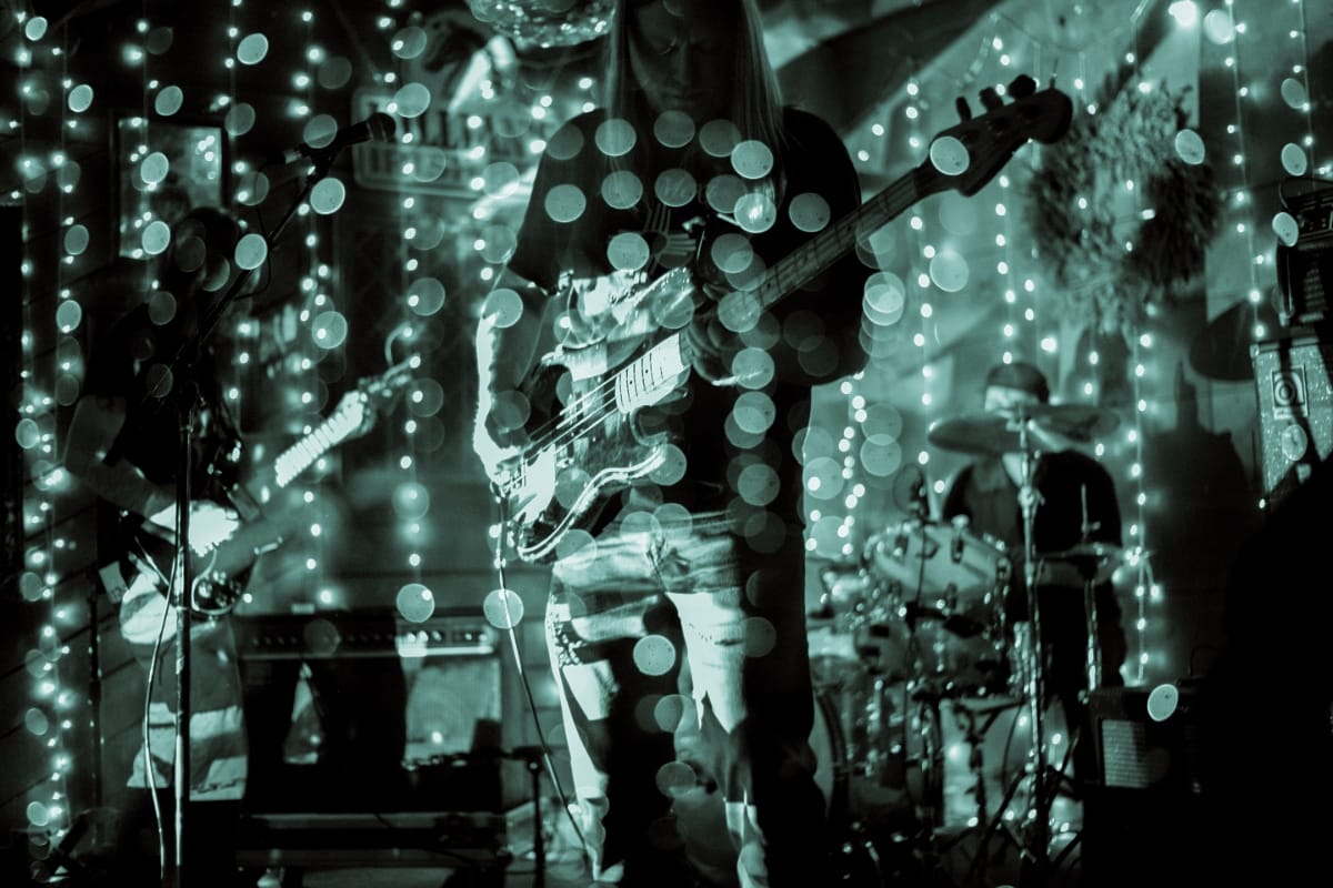 Unwed Sailor perform on a small stage strung with fairy lights, a bassist centered in foreground, guitarist left, drummer right, all cast in teal-tinted light with heavy bokeh. Photo by Alejandro Martinez.