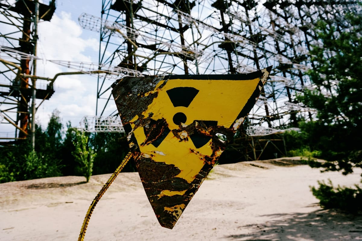 A weathered, rust-streaked radiation warning sign in yellow and black hangs in the foreground, with the massive steel lattice structure of the Duga radar array rising behind it amid green trees.