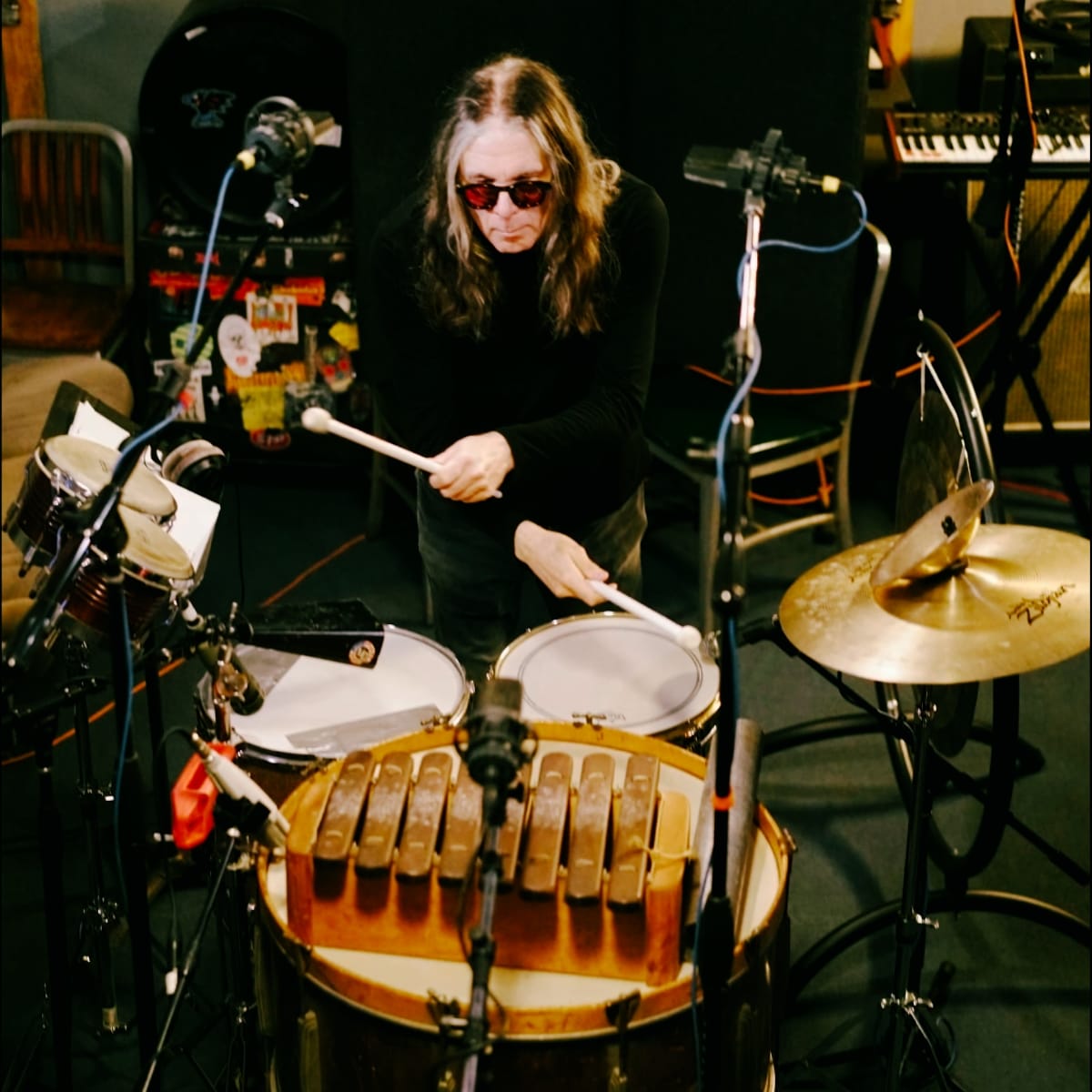 Bob Bert photographed from above, playing snare and xylophone surrounded by mic stands and cymbals, wearing a black turtleneck and red-tinted sunglasses. Credit: Mark C.