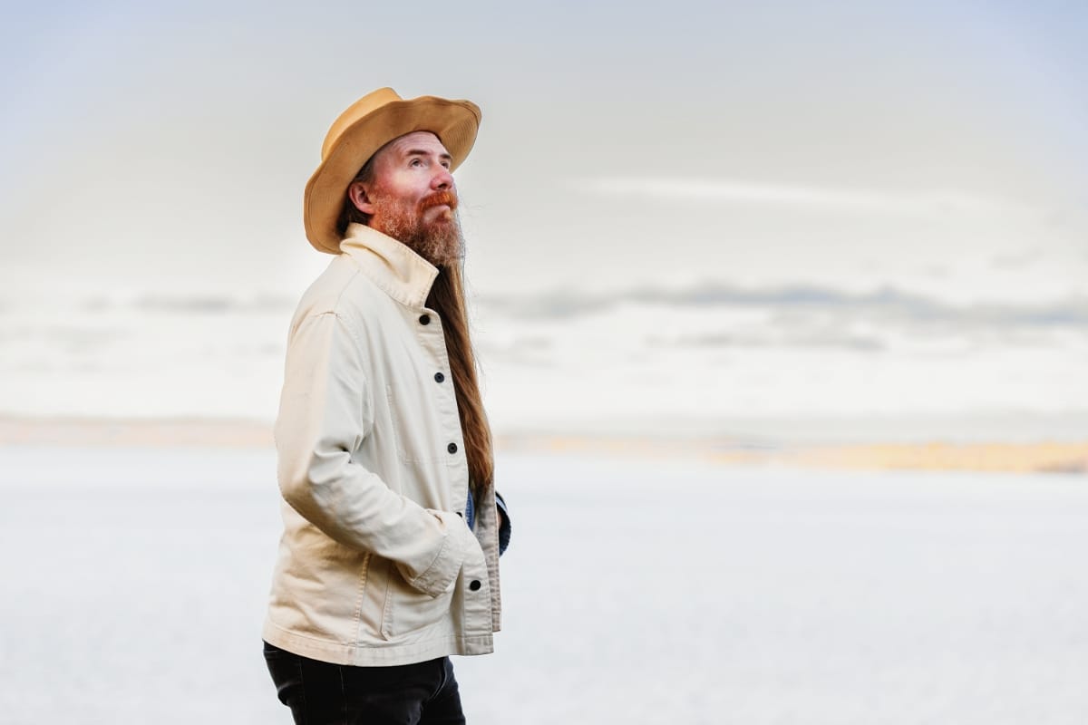 Bob Wagner stands in profile against a pale open landscape, long auburn hair and beard, tan wide-brim hat, cream jacket, gazing upward.