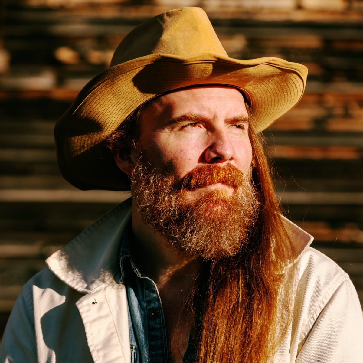 Bob Wagner, wide-brimmed tan hat and long auburn hair and beard, looks upward against a warm-lit wooden wall.