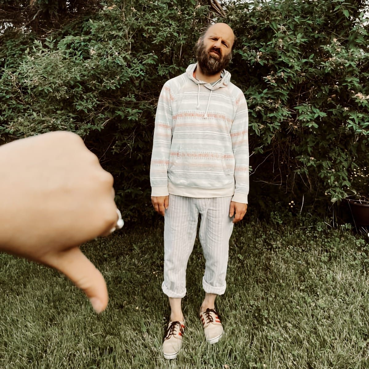 Chik White stands in a backyard, bearded and bald, wearing a striped hoodie and cuffed trousers, as a hand in the foreground gives a thumbs-down.