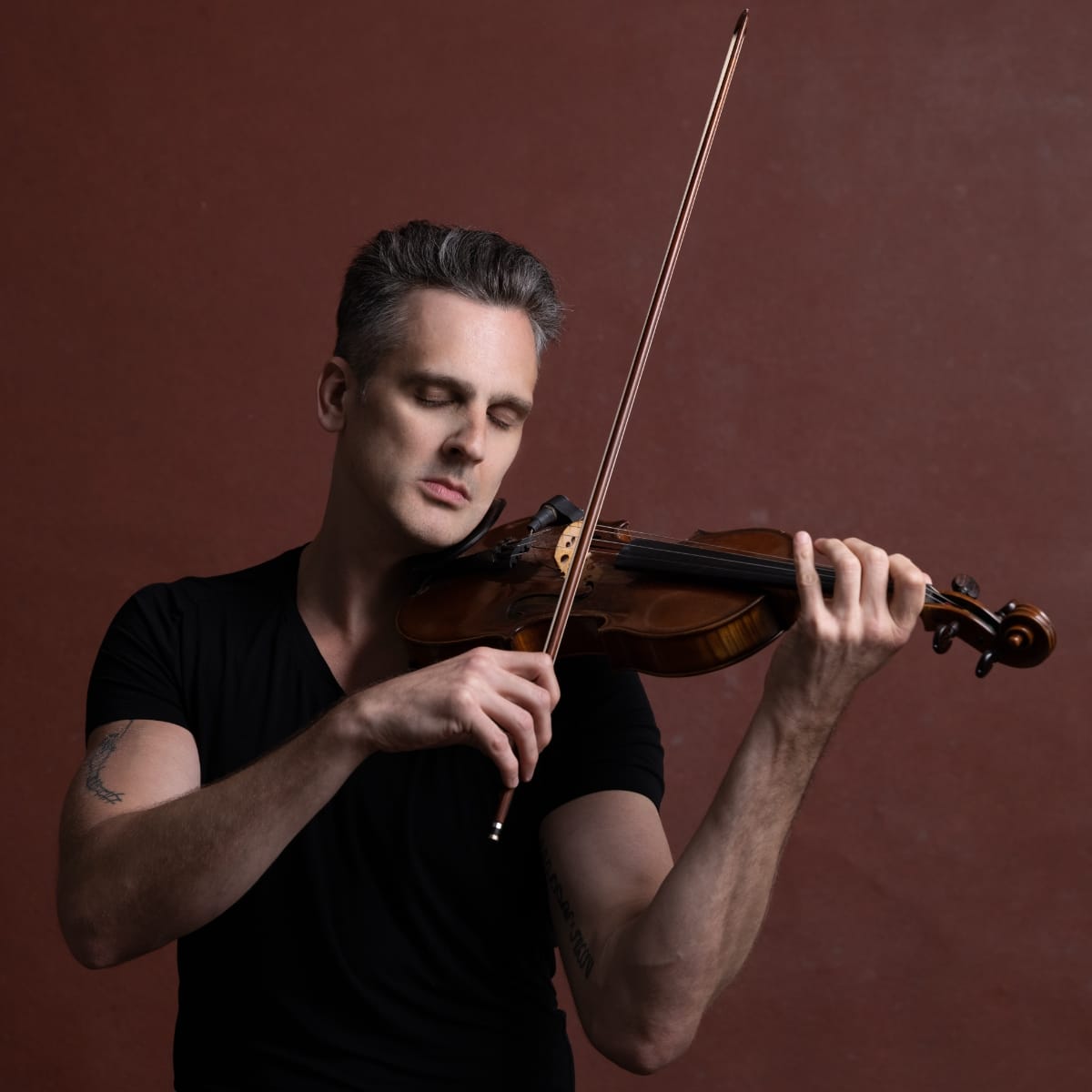 Christopher Tignor plays violin against a deep red-brown backdrop, eyes closed, bow raised. A tattoo is visible on his left arm. A small clip-on mic is attached near the instrument's chin rest.