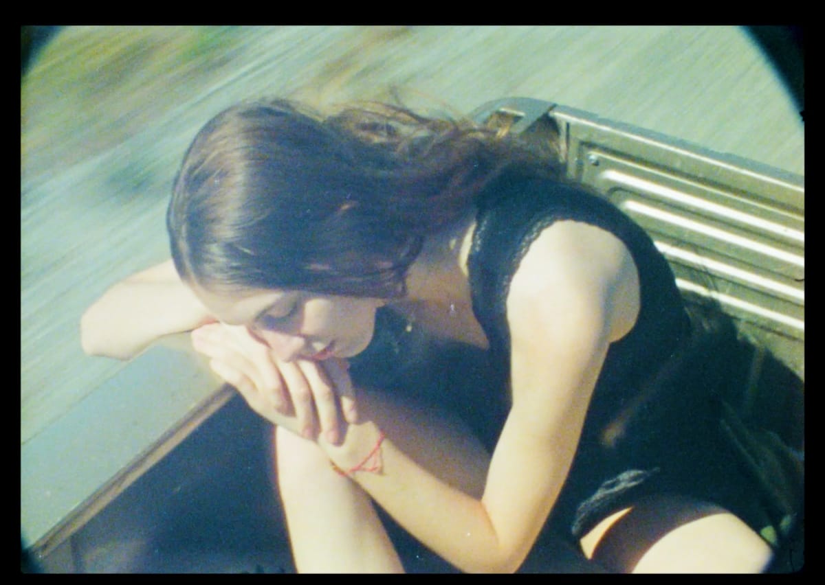 Eliana Glass sits in what appears to be a pickup truck bed, head bowed, arms folded over her knees. Shot from above on grainy film stock with a vignetted border.