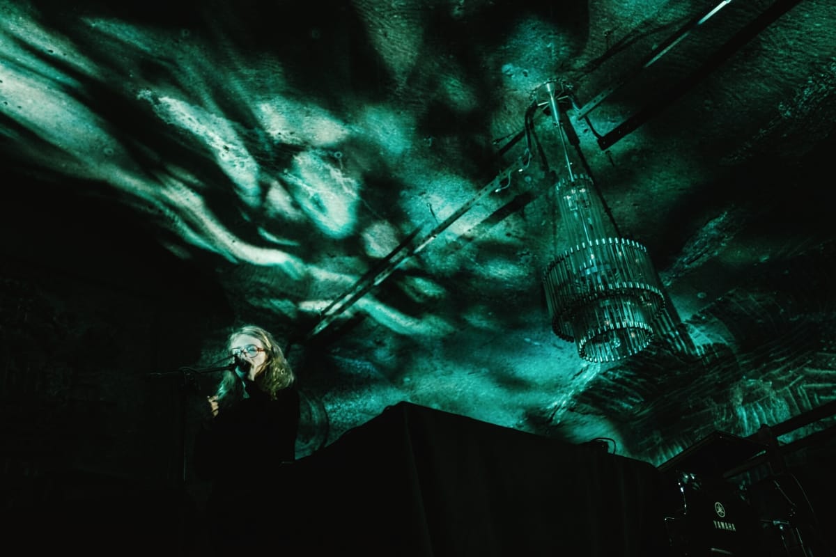 Félicia Atkinson performs at a keyboard in near-darkness, a microphone at her lips, while teal-lit projections of fluid shadows sweep across the ceiling above a hanging chandelier.