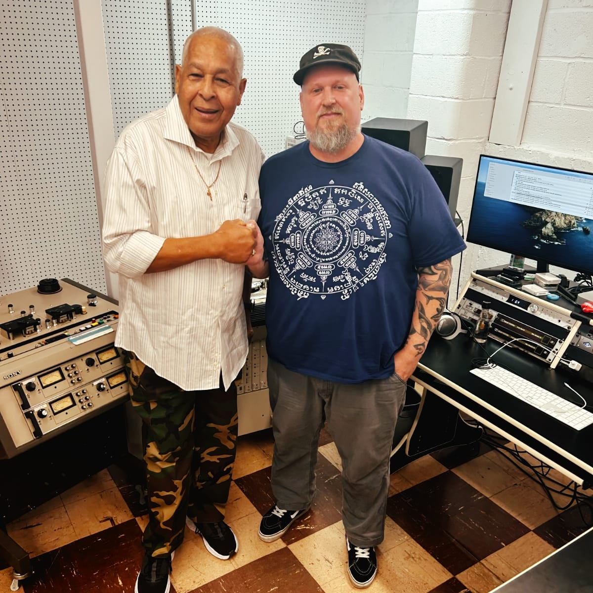 King Jammy and Billy Polo stand together in a recording studio, a reel-to-reel tape machine visible to their left and studio gear along the wall behind them.