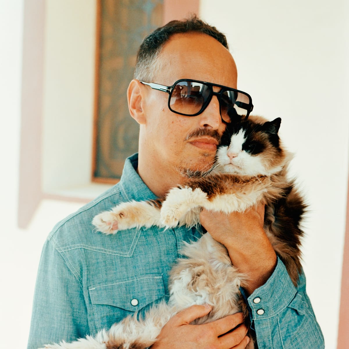 Mark Barrott holds a long-haired calico cat pressed against his cheek, both facing the camera, against a bright interior wall.