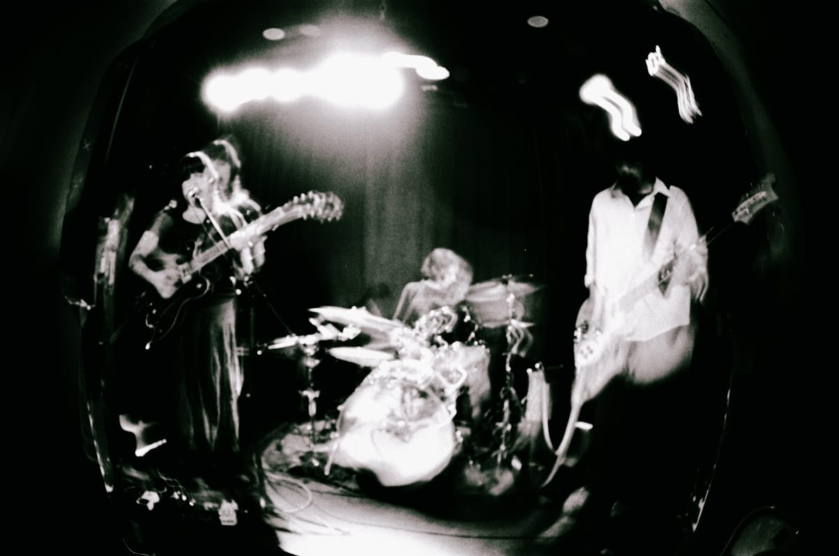 Myra Lee perform live in a grainy black-and-white fisheye shot, motion blur streaking the edges as guitarist, drummer, and bassist fill a spotlight-lit stage.