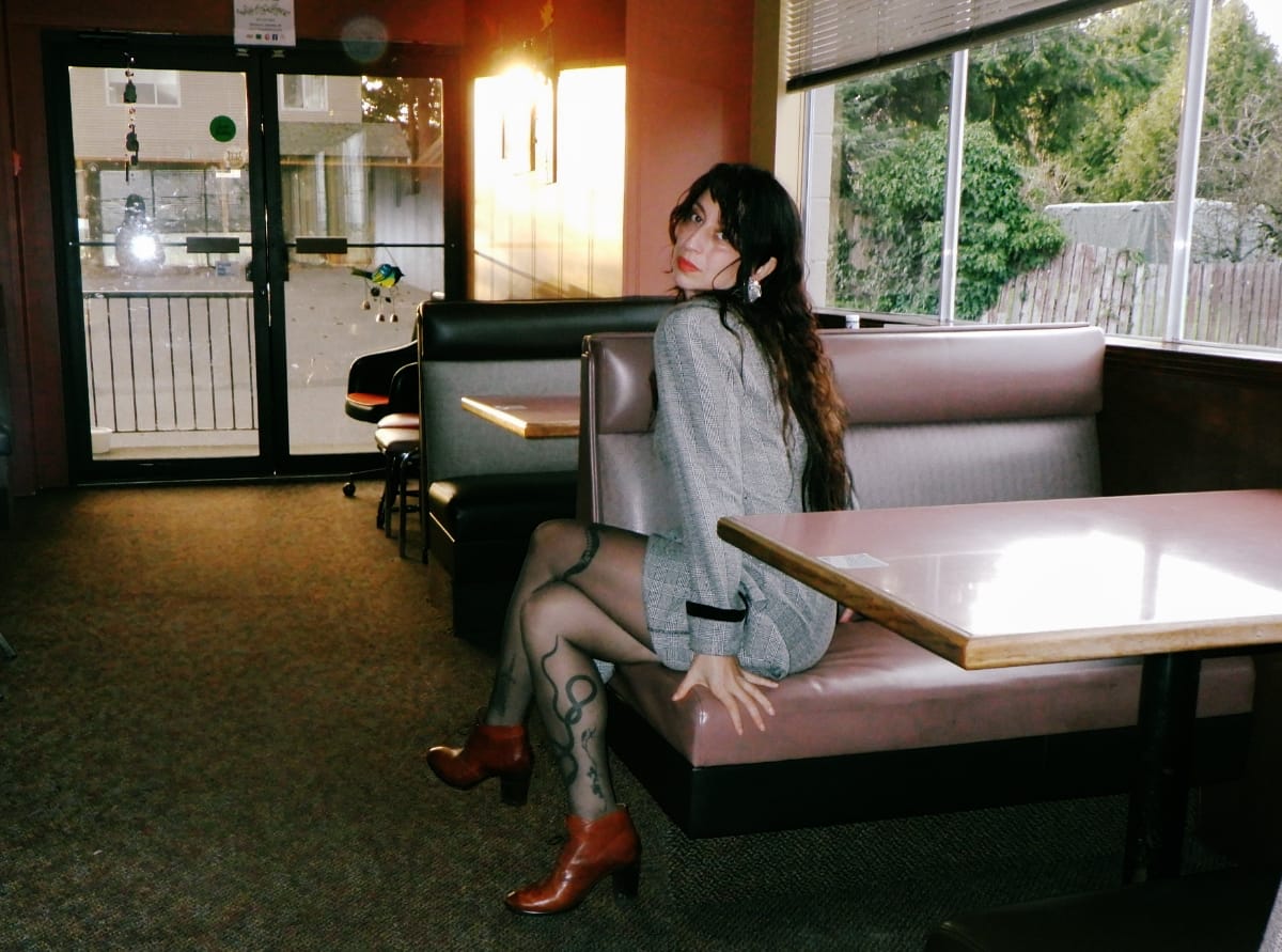 Ora Cogan sits in a diner booth, glancing back at the camera, long dark hair loose, wearing a gray blazer dress, snake-patterned tights, and rust-colored ankle boots.