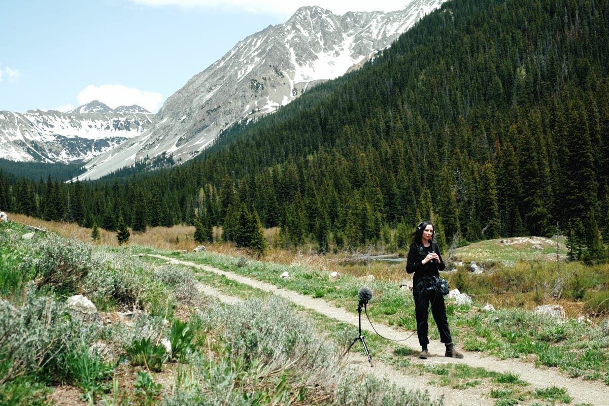 Patricia Wolf stands on a dirt path with headphones on and a recorder in hand, a boom mic on a tripod beside her, snow-capped mountains and pine forest stretching behind her.