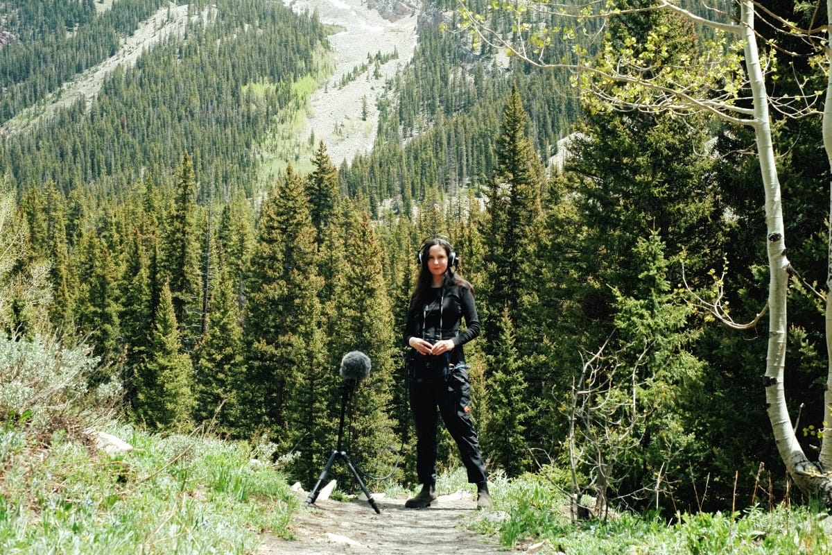 Patricia Wolf stands on a mountain trail with headphones on, a boom microphone on a tripod beside her, dense evergreen forest and a rocky slope rising behind her.