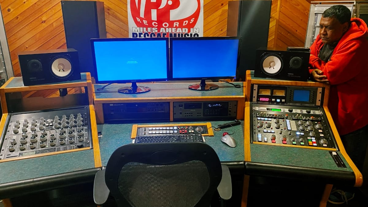 Paul Shields stands at the edge of the VP Records mastering room, a large console flanked by studio monitors and dual screens filling the foreground, a VP Records banner on the wall behind.