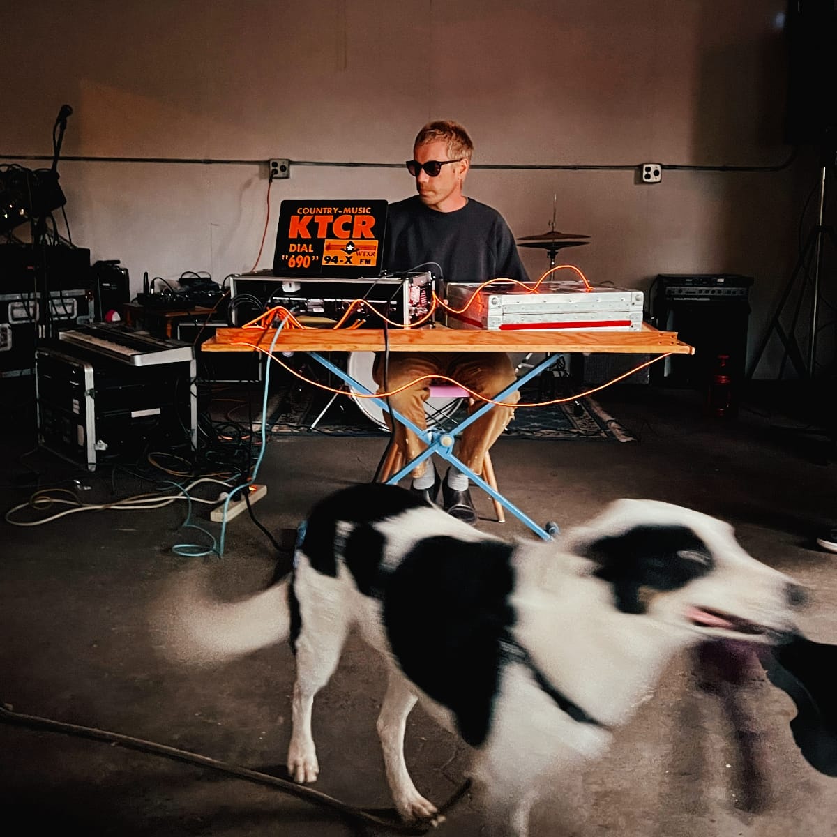 Phil Geraldi performs behind a folding table covered in gear and orange patch cables in a bare room. A black-and-white dog roams the foreground. A vintage KTCR Country Music ticker in on his laptop.