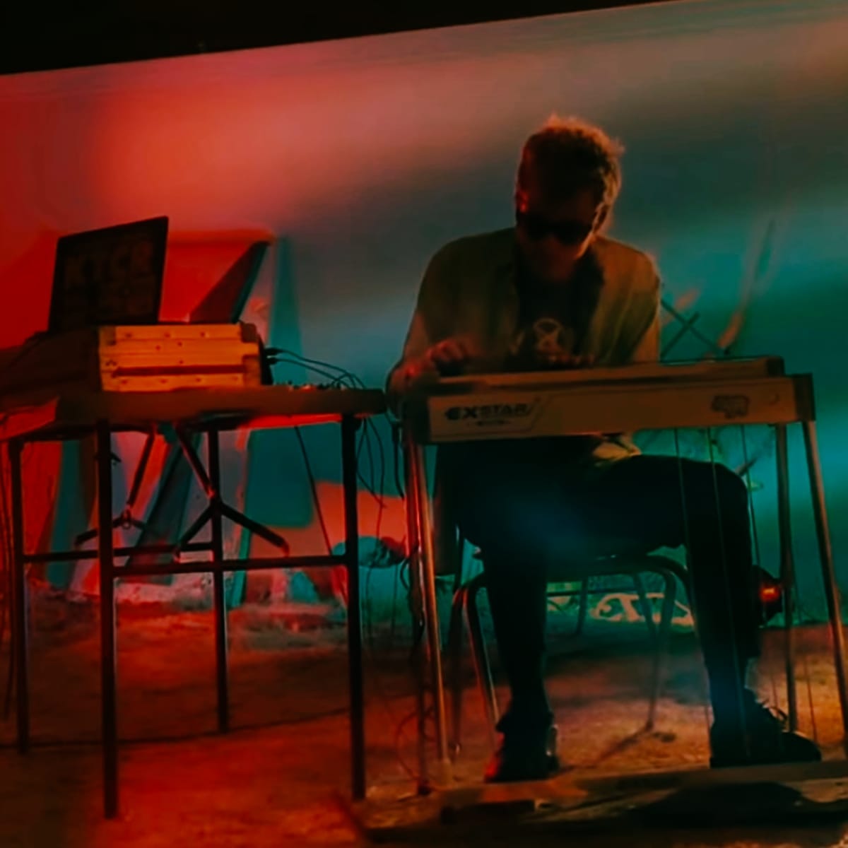 Phil Geraldi performs seated at a table of electronic gear, bathed in deep red and teal lighting. A laptop and equipment cases sit to his left against a painted backdrop.