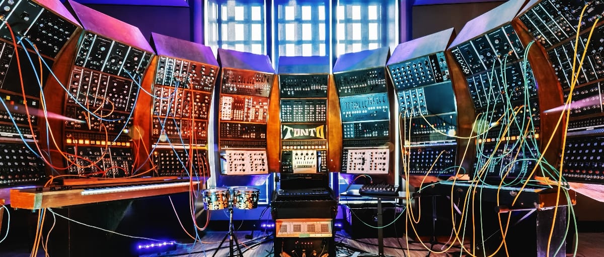 TONTO, a massive curved wall of modular synthesizer panels, fills the frame floor to ceiling, dozens of multicolored patch cables radiating outward from its banks of knobs and modules.
