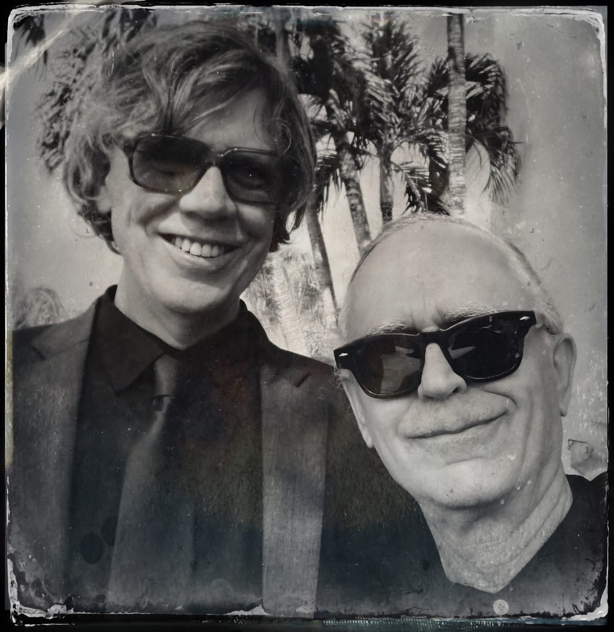 Thurston Moore and Bonner Kramer smile for a tintype-style black-and-white photo outdoors, both in sunglasses, palm trees behind them.