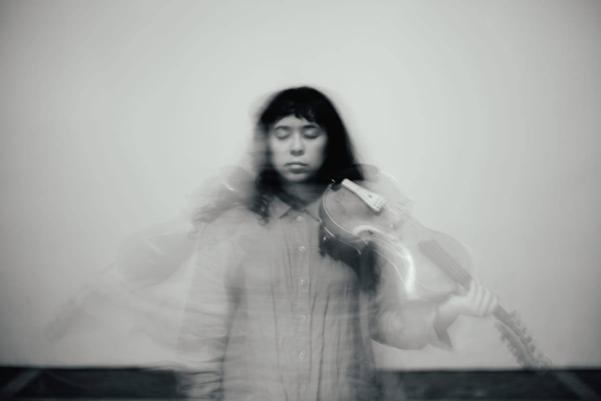 Zosha Warpeha holds a violin against her shoulder, eyes closed, in a black-and-white photo shot with heavy motion blur that dissolves her figure into soft ghostly streaks.