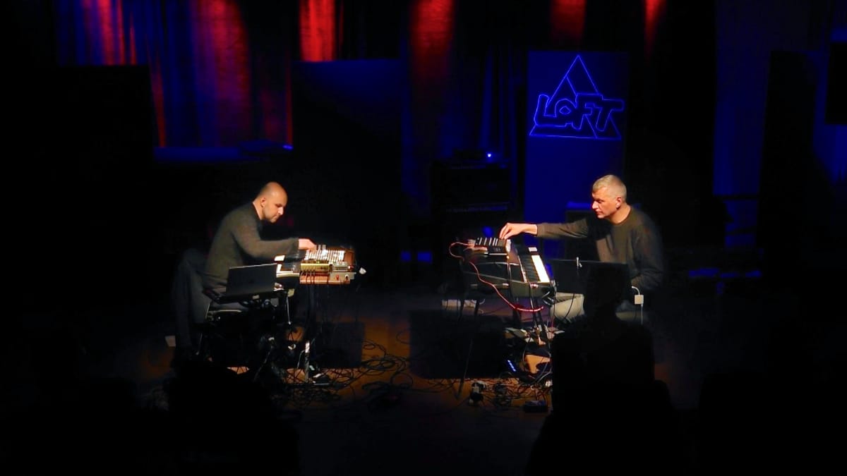 Joseph Branciforte and Jozef Dumoulin perform at The Loft, Cologne, each bent over a keyboard and electronics setup on a darkened stage lit in red and blue.