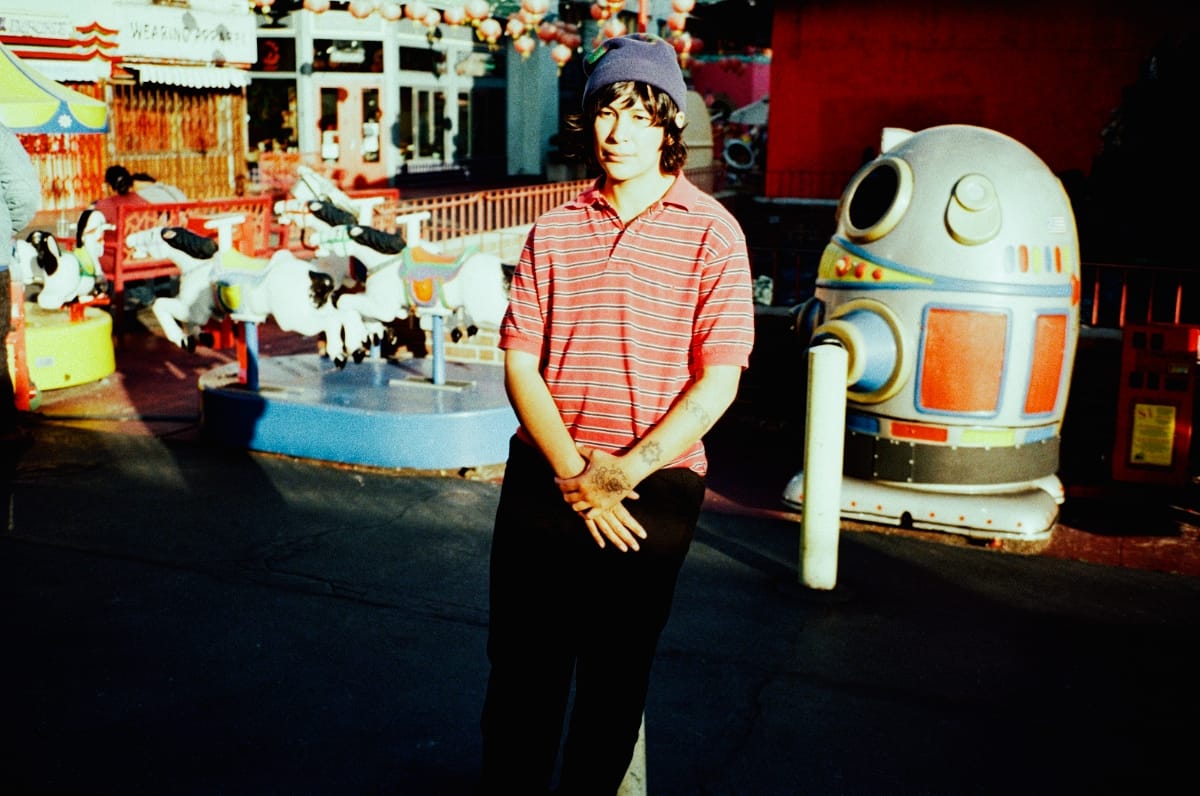 zzzahara standing at an amusement park, hands clasped, wearing a blue beanie and red striped polo. A carousel and a retro robot ride machine flank them in the background. Photo by Lindsey Byrnes.